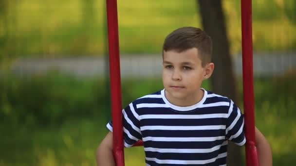 Een jongetje in een gestreepte T-shirt is het spelen op de speelplaats, Swing op een schommel. Lente, zonnig weer. — Stockvideo