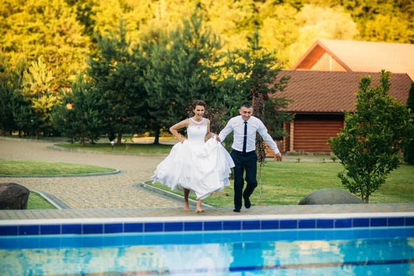 Jong (echt) paar springen in het zwembad in een bruiloft pak en bruiloft jurk. Zonnige dag. — Stockfoto