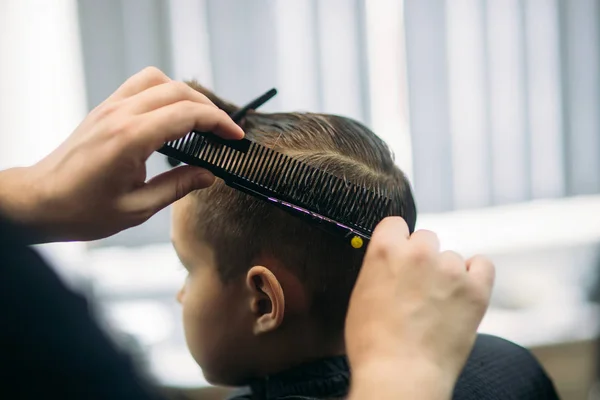 Pequeño chico consiguiendo corte de pelo por peluquero mientras está sentado en silla en la barbería . — Foto de Stock