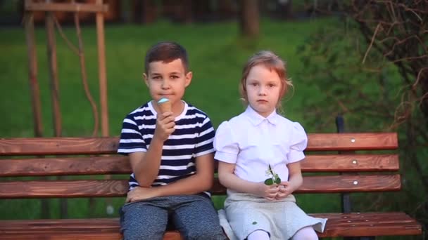 Хлопчик і дівчинка сидять на лавці. Бігати навколо парку і дути кульбаби — стокове відео