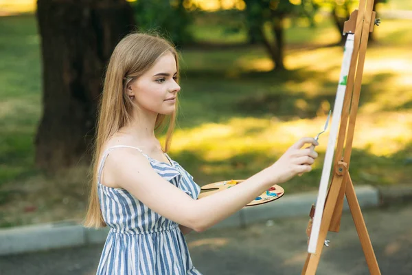 Blond dziewczyna farby malowanie na płótnie przy użyciu farb. Drewniane sztalugi trzyma obraz. — Zdjęcie stockowe