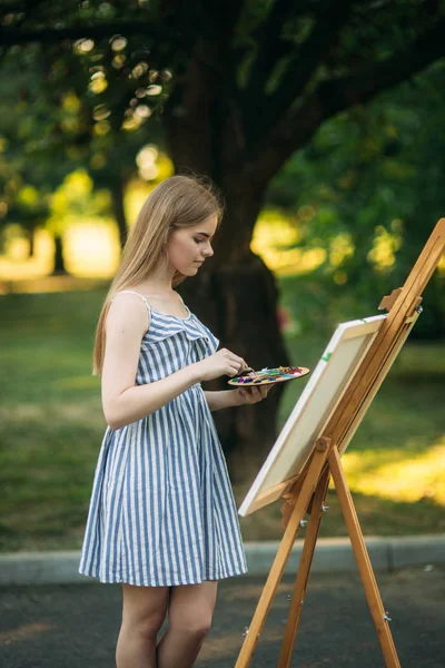 La chica rubia pinta un cuadro en el lienzo con la ayuda de pinturas. Un caballete de madera mantiene la imagen . — Foto de Stock