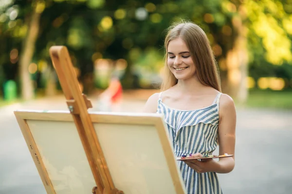 Het blonde meisje schildert een schilderij op het canvas met de hulp van verven. Een houten ezel houdt de afbeelding. — Stockfoto