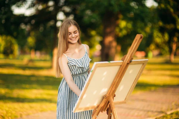 Όμορφο κορίτσι τραβάει μια εικόνα στο πάρκο χρησιμοποιώντας μια παλέτα με χρώματα και μια σπάτουλα. Καβαλέτο και καμβάς με την εικόνα. Το καλοκαίρι είναι μια ηλιόλουστη μέρα, ηλιοβασίλεμα. — Φωτογραφία Αρχείου