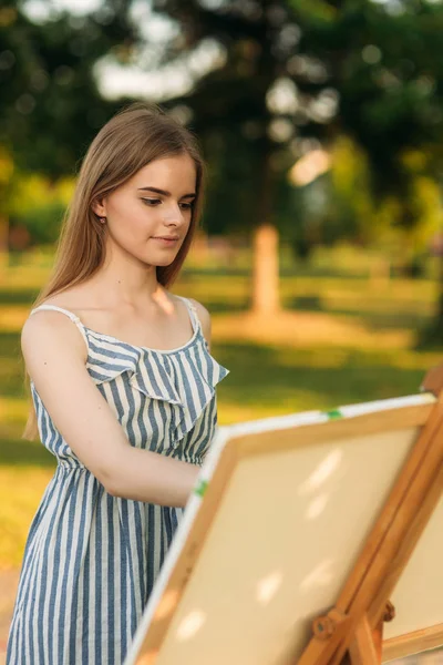 Hermosa chica dibuja un cuadro en el parque usando una paleta con pinturas y una espátula. caballete y lienzo con una imagen. El verano es un día soleado, puesta de sol . — Foto de Stock