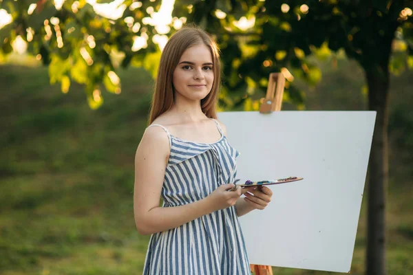 Piękna dziewczyna rysuje obraz w parku przy użyciu palety farb i łopatką. Sztalugi i płótno z rysunkiem. Lato jest słoneczny dzień, zachód słońca. — Zdjęcie stockowe
