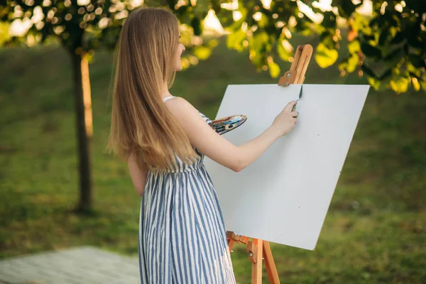 Menina bonita desenha um quadro no parque usando uma paleta com tintas e uma espátula. Easel e lona com uma foto. O verão é um dia ensolarado, pôr do sol . — Fotografia de Stock
