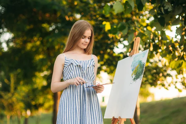 漂亮的女孩在公园里使用一个调色板，用油漆和一把铲子画一幅画。画架和画布上用一张图片。夏天是一个阳光明媚的日子，日落. — 图库照片