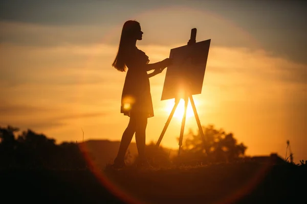 A silhueta de uma rapariga. A menina loira pinta uma pintura na tela com a ajuda de tintas. Um cavalete de madeira mantém a imagem . — Fotografia de Stock
