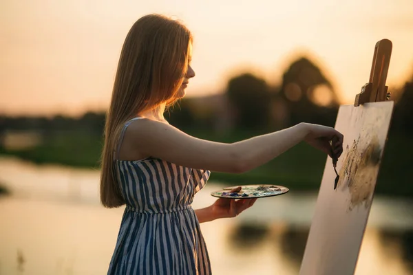 Hermosa chica dibuja un cuadro en el parque usando una paleta con pinturas y una espátula. caballete y lienzo con una imagen. El verano es un día soleado, puesta de sol . — Foto de Stock