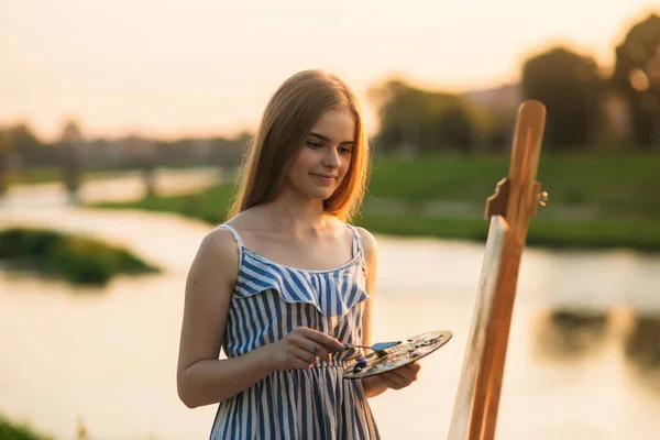 Hermosa chica dibuja un cuadro en el parque usando una paleta con pinturas y una espátula. caballete y lienzo con una imagen. El verano es un día soleado, puesta de sol . — Foto de Stock