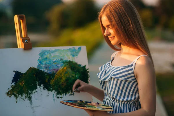 Piękna dziewczyna rysuje obraz w parku przy użyciu palety farb i łopatką. Sztalugi i płótno z rysunkiem. Lato jest słoneczny dzień, zachód słońca. — Zdjęcie stockowe