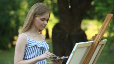 Güzel kız parkta boyalar ve bir spatula ile bir palet kullanarak bir resim çizer. Şövale ve tuval resmi.