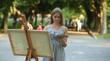 Güzel kız parkta boyalar ve bir spatula ile bir palet kullanarak bir resim çizer. Şövale ve tuval resmi.