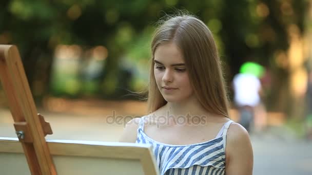 Hermosa chica dibuja un cuadro en el parque usando una paleta con pinturas y una espátula. caballete y lienzo con una imagen . — Vídeo de stock