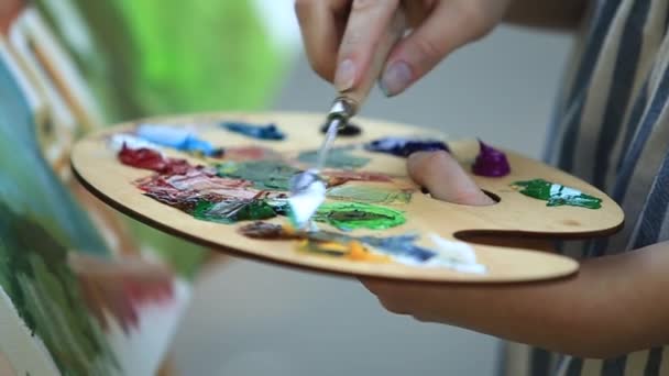 Beautiful girl draws a picture in the park using a palette with paints and a spatula. Easel and canvas with a picture. — Stock Video