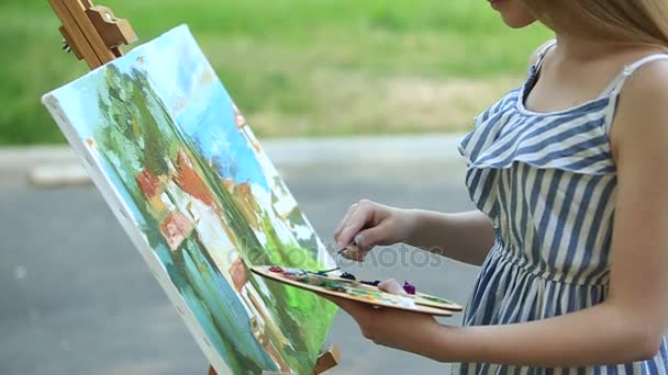 Hermosa chica dibuja un cuadro en el parque usando una paleta con pinturas y una espátula. caballete y lienzo con una imagen . — Vídeos de Stock