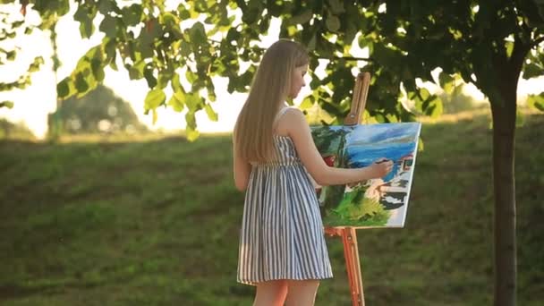 Menina bonita desenha um quadro no parque usando uma paleta com tintas e uma espátula. Easel e lona com uma imagem . — Vídeo de Stock