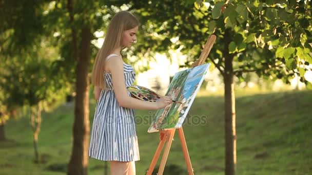 Bella ragazza disegna un disegno nel parco utilizzando una tavolozza con vernici e una spatola. Cavalletto e tela con un'immagine . — Video Stock