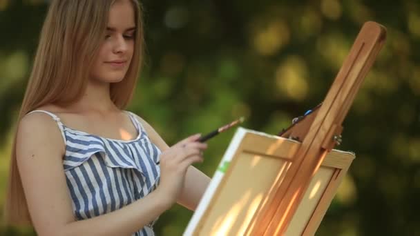 Belle fille dessine une image dans le parc en utilisant une palette avec des peintures et une spatule. Chevalet et toile avec une image . — Video