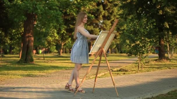Όμορφο κορίτσι τραβάει μια εικόνα στο πάρκο χρησιμοποιώντας μια παλέτα με χρώματα και μια σπάτουλα. Καβαλέτο και καμβάς με την εικόνα. — Αρχείο Βίντεο
