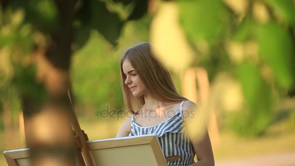 Hermosa chica dibuja un cuadro en el parque usando una paleta con pinturas y una espátula. caballete y lienzo con una imagen . — Vídeos de Stock