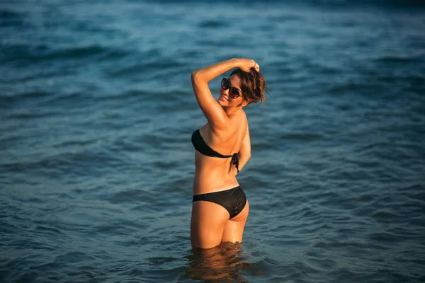 Sexy chica tomando el sol en una playa de arena y se sienta en la orilla del mar —  Fotos de Stock