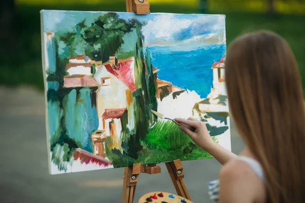 Güzel kız parkta boyalar ve bir spatula ile bir palet kullanarak bir resim çizer. Şövale ve tuval resmi.