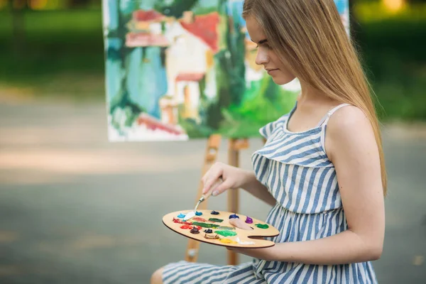 Mooi meisje tekent een afbeelding in het park met behulp van een palet met verf en een spatel. Ezel en canvas met een afbeelding. — Stockfoto