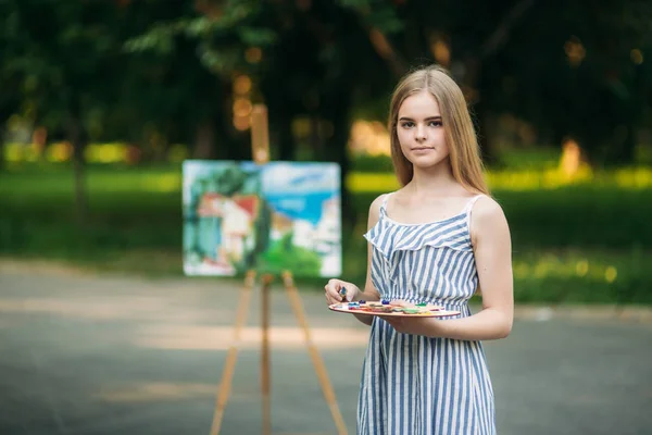 Mooi meisje tekent een afbeelding in het park met behulp van een palet met verf en een spatel. Ezel en canvas met een afbeelding. — Stockfoto