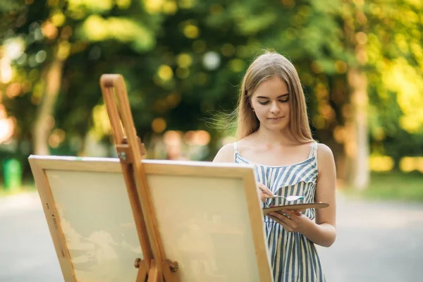 Красива дівчина малює в парку фотографію, використовуючи палітру з фарбами і шпателем. Мольберт і полотно з малюнком . — стокове фото