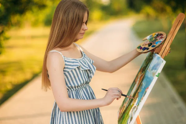 Красива дівчина малює в парку фотографію, використовуючи палітру з фарбами і шпателем. Мольберт і полотно з малюнком . — стокове фото