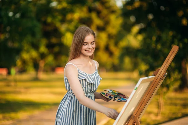 Hermosa chica dibuja un cuadro en el parque usando una paleta con pinturas y una espátula. caballete y lienzo con una imagen . — Foto de Stock
