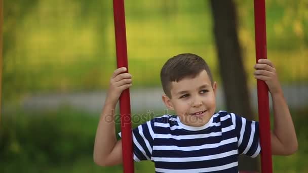 Un niño pequeño con una camiseta a rayas está jugando en el patio de recreo, Swing on a swing.Spring . — Vídeo de stock