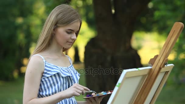 Hermosa chica dibuja un cuadro en el parque usando una paleta con pinturas y una espátula. caballete y lienzo con una imagen. El verano es un día soleado, puesta de sol . — Vídeo de stock