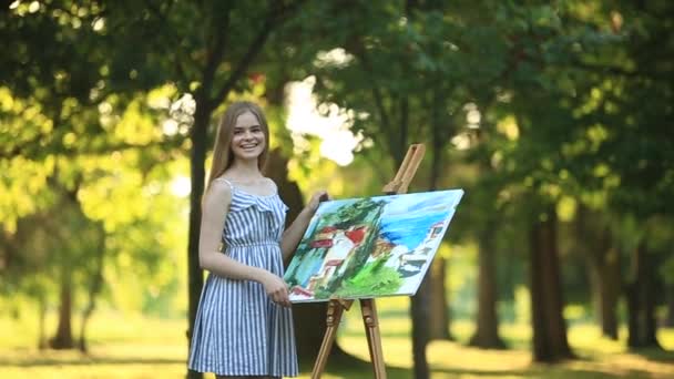 Mooi meisje tekent een afbeelding in het park met behulp van een palet met verf en een spatel. Ezel en canvas met een afbeelding. De zomer is een zonnige dag, zonsondergang. — Stockvideo