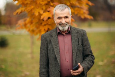 Gözlük takan yakışıklı bir yaşlı adam bir telefon kullanıyor. Sonbaharda Parkta yürümek