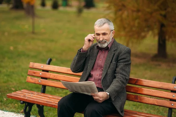 Un bel nonno con una bella barba in giacca grigia siede su una panchina nel parco e legge un giornale. — Foto Stock