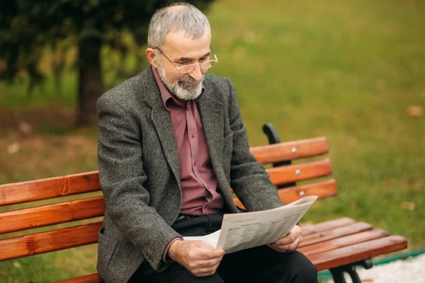 Хороший дедушка с красивой бородой в серой куртке сидит на скамейке в парке и читает газету — стоковое фото