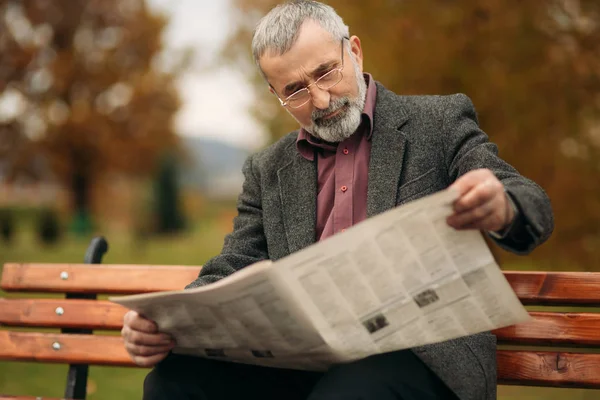 Un buen abuelo con una hermosa barba en una chaqueta gris se sienta en un banco en el parque y lee un periódico —  Fotos de Stock