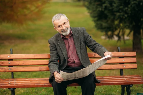 Un buen abuelo con una hermosa barba en una chaqueta gris se sienta en un banco en el parque y lee un periódico —  Fotos de Stock