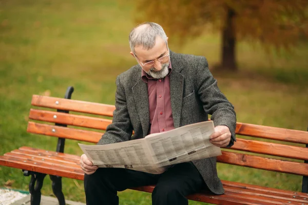 Un gentil grand-père avec une belle barbe dans une veste grise s'assoit sur un banc dans le parc et lit un journal — Photo