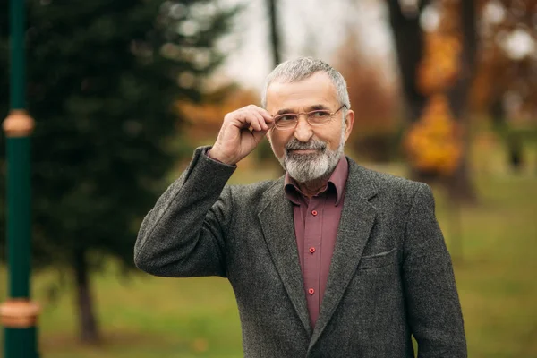 Un bell'uomo anziano con gli occhiali sta usando un telefono. Passeggiata nel parco in autunno — Foto Stock