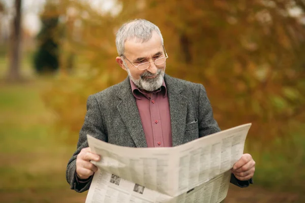 Un buen abuelo con una hermosa barba en una chaqueta gris se sienta en un banco en el parque y lee un periódico —  Fotos de Stock