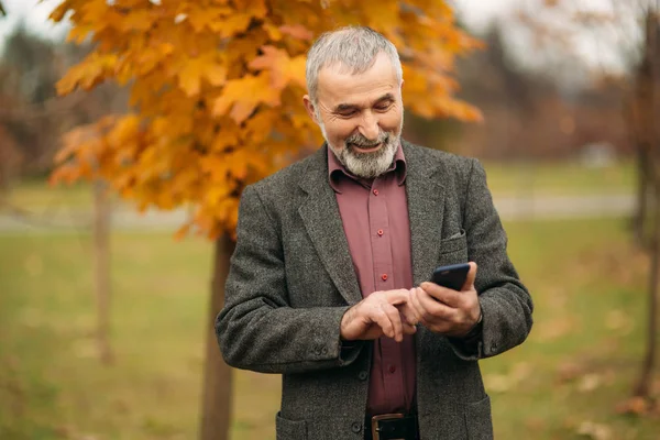En stilig äldre man som bär glasögon använder en telefon. Promenad i parken under hösten — Stockfoto