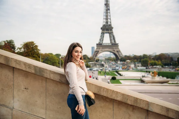 Beautiful girl posing to the photographer against the background of the Eiffel Tower. Autumn photosession. Sunny weather. Beautiful smile and makeup