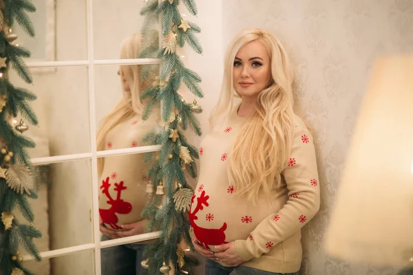 En ganska gravid blondin i jul tröja står nära spegeln. — Stockfoto