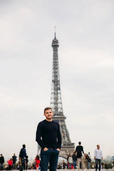 Un joven con una chaqueta azul oscuro está de pie en el fondo de la Torre Eiffel. El tiempo soleado es otoño — Foto de Stock