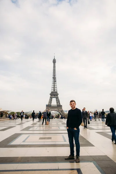 Um jovem vestindo um casaco azul escuro está no fundo da Torre Eiffel. Tempo ensolarado é outono — Fotografia de Stock