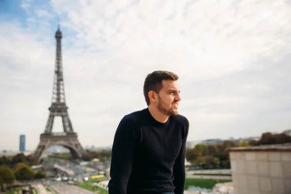 Un joven con una chaqueta azul oscuro está de pie en el fondo de la Torre Eiffel. El tiempo soleado es otoño — Foto de Stock